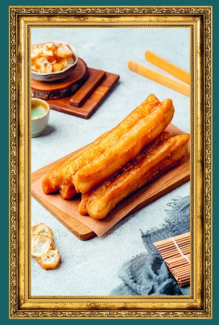 Ein gerahmtes Foto von knusprigen chinesischen Youtiao (frittierten Teigstäbchen), die auf einem Holzbrett gestapelt sind, mit Dip-Sauce und geschnittenen Stücken im Hintergrund auf einer hellen Fläche.