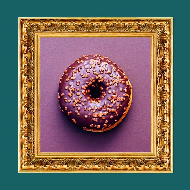 Ein Donut mit Schokoladenglasur und Streuseln wird vor einem violetten Hintergrund zentriert und in einem verschnörkelten, goldenen Bilderrahmen präsentiert, wodurch eine kunstvolle, galerieähnliche Präsentation entsteht.