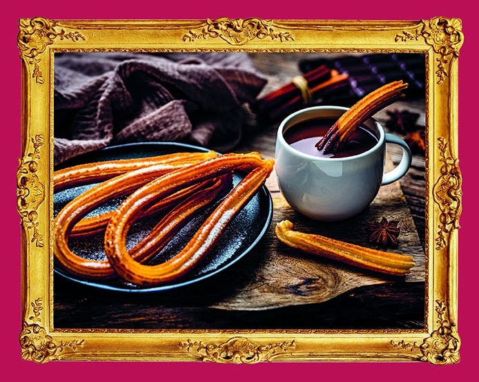 Goldene, mit Zucker überzogene Churros auf einem schwarzen Teller neben einer weißen Tasse mit heißer Schokolade, wobei ein Churro in die Tasse getaucht ist. Das Foto ist in einem verschnörkelten Goldrahmen vor einem magentafarbenen Hintergrund gerahmt.