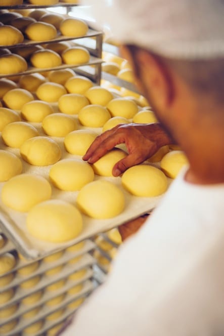 Eine Person stellt ein Tablett mit ordentlich angeordneten, runden gelben Teigkugeln auf ein Gestell, das mit ähnlichen Tabletts in einer Bäckerei gefüllt ist.