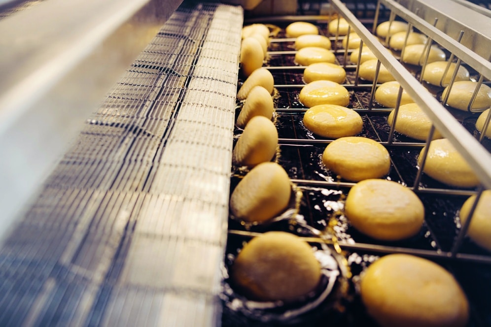 Reihen von runden Teiglingen, die auf einem industriellen Fließband in Öl frittiert werden, wahrscheinlich in einer Fabrik für die Massenproduktion von Donuts oder ähnlichen Backwaren.