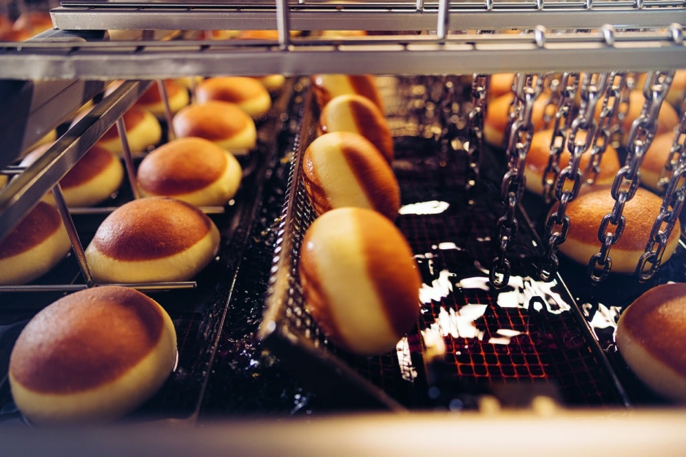 Nahaufnahme von runden Krapfen, die auf einem Förderband in einer Bäckerei oder Fabrik in Öl frittiert werden.