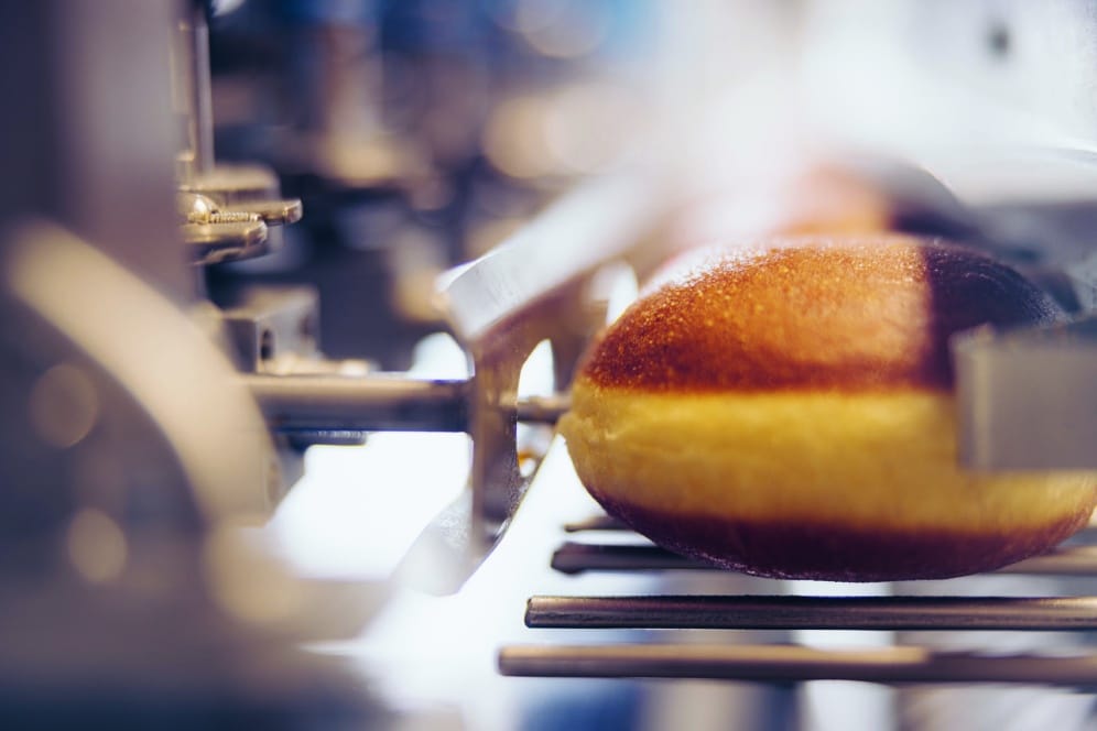 Nahaufnahme eines goldbraunen Gebäcks oder Doughnuts, das auf einem Metallförderband in einer Lebensmittelverarbeitungs- oder -herstellungsanlage entlangläuft, wobei im Hintergrund Maschinen zu sehen sind.