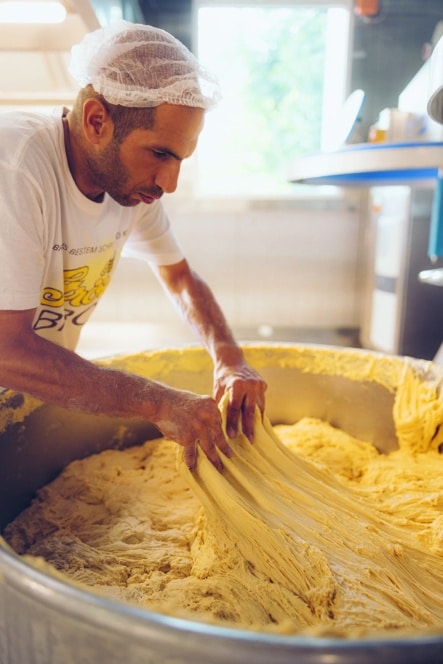 Ein Mann mit Haarnetz und weißem T-Shirt knetet oder streckt eine große Menge gelben Teigs in einem Metallbehälter in einer hellen, modernen Küche oder Bäckerei.