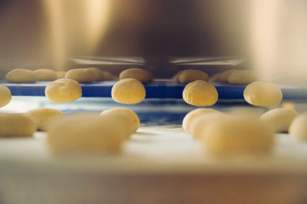 Nahaufnahme von Reihen kleiner, runder Teigkugeln auf einem Förderband in einer Bäckerei oder Fabrik, mit geringer Schärfentiefe, die den Teig im Vordergrund betont.