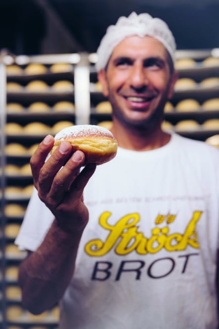 Ein lächelnder Bäcker mit Haarnetz und weißem Hemd mit der Aufschrift "Brot" hält einen mit Marmelade gefüllten Donut in einer Bäckerei hoch, während im Hintergrund Tabletts mit Brot stehen.