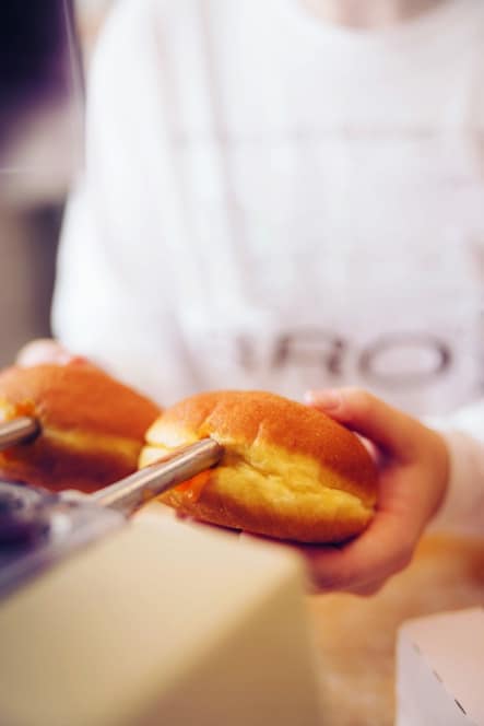 Eine Person hebt mit einer Metallzange ein rundes, goldbraunes Gebäck oder einen Krapfen auf und hält in der anderen Hand ein anderes Gebäck. Der Hintergrund ist sanft verschwommen.