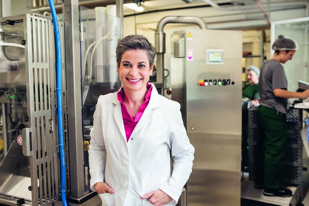 Eine Frau in einem weißen Laborkittel und einem rosa Hemd lächelt, während sie in einem Labor oder einer Industrieanlage steht, in der im Hintergrund Maschinen und zwei andere Personen arbeiten.
