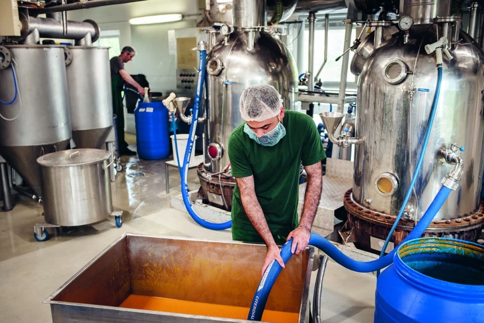 Ein Arbeiter in grünem Hemd und Haarnetz füllt ein blaues Fass mit Flüssigkeit aus einem Edelstahlbehälter in einer Fabrik, während ein anderer Arbeiter im Hintergrund in der Nähe von Industrieanlagen steht.