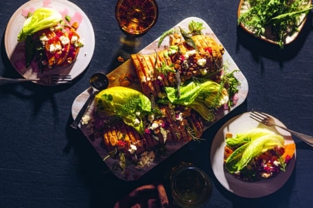 Draufsicht auf einen Tisch mit Tellern mit bunten Tacos, gefüllt mit Gemüse, Bohnen, Käse und frischem Salat, umgeben von Getränken und einer Schale mit Grünzeug auf einer dunklen Fläche.
