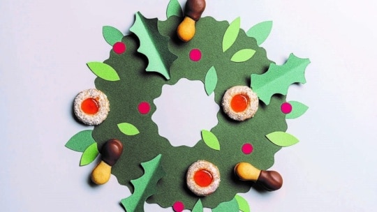 Ein grüner Papierkranz mit Stechpalmenblättern, roten Beeren und verschiedenen Keksen, darunter mit Gelee gefüllte und in Schokolade getauchte Leckereien, auf einem hellen Hintergrund.