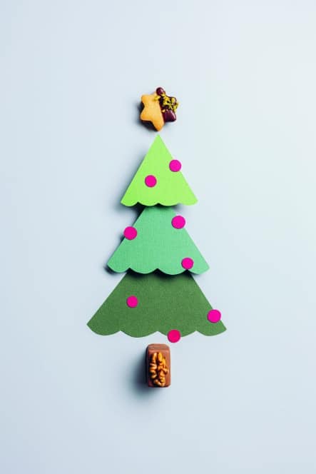 Ein minimalistischer Weihnachtsbaum aus drei grünen Papierausschnitten mit rosa Punkten, gekrönt von einem Lebkuchenstern und einem kleinen braunen Gebäckstück an der Basis, angeordnet auf einem hellen Hintergrund.