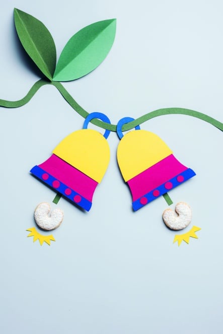 Zwei bunte Papierglocken mit grünen Stielen und Blättern, verziert mit rosa und blauen Bändern, hängen nebeneinander. An jeder Glocke baumelt ein Keksherz als Klöppel vor einem hellblauen Hintergrund.