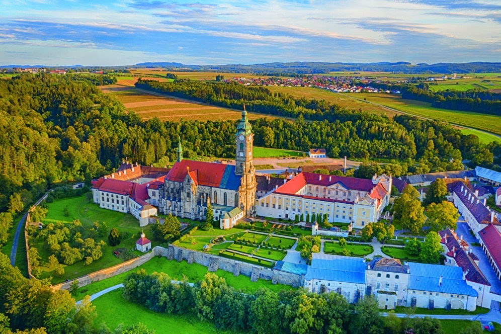 Luftaufnahme eines großen historischen Klosterkomplexes mit roten Dächern, einem zentralen Uhrenturm und gepflegten Gärten, umgeben von grünen Bäumen und Feldern unter einem teilweise bewölkten Himmel.
