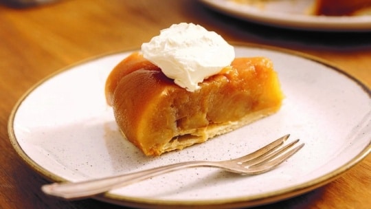 Ein Stück Tarte Tatin mit einem Klecks Schlagsahne liegt auf einem weißen Teller mit einer silbernen Gabel, der auf einem Holztisch steht.