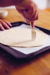 Eine Hand sticht mit einer Gabel Löcher in ein rundes Blatt rohen Teigs, das auf einem mit Pergamentpapier ausgelegten Backblech liegt, um es für das Backen vorzubereiten.