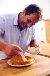 Ein Mann in einem hellblauen Hemd lächelt, während er in einer hellen Küche einen runden Kuchen auf einem Teller mit Glasur bestreicht und in der anderen Hand eine kleine Schüssel hält.
