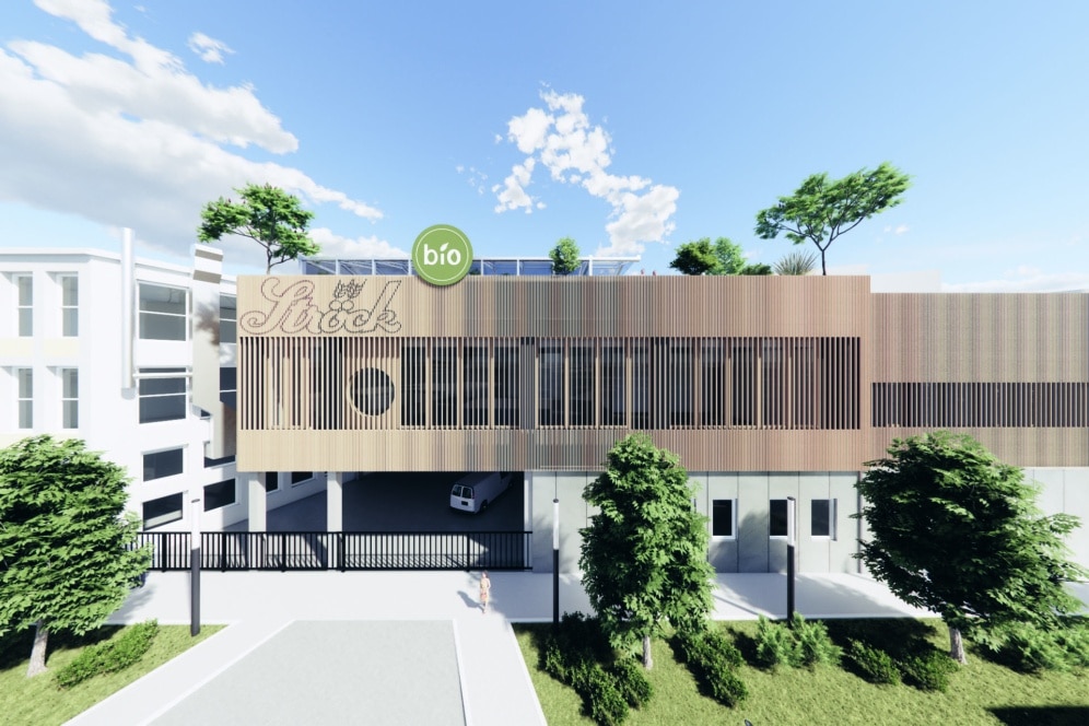 Ein modernes Gebäude mit hellbrauner Fassade und vertikalen Lamellen, auf denen ein grünes, rundes "Bio"-Schild und das "Ströck"-Logo angebracht sind. Auf dem Dach und um das Gebäude herum sind Bäume und Grünflächen zu sehen, über denen ein strahlend blauer Himmel liegt.