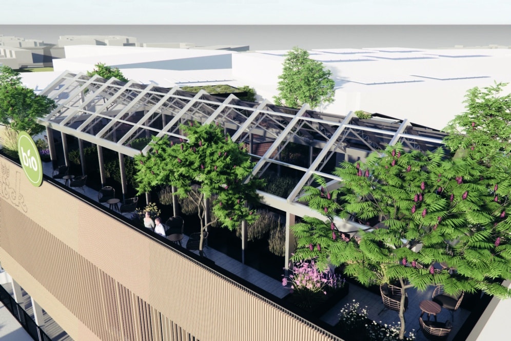 Eine Dachterrasse mit einer gläsernen Pergolastruktur, üppig grünen Bäumen und Topfpflanzen. Unter der Pergola sind mehrere Tische und Stühle aufgestellt, die einen schattigen Sitzbereich im Freien bilden. Im Hintergrund sind städtische Gebäude zu sehen.