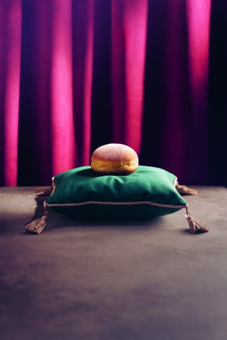 Ein einzelner gepuderter Donut sitzt auf einem plüschigen grünen Kissen mit goldenen Quasten vor einer dunklen Oberfläche und sattvioletten Vorhängen im Hintergrund.