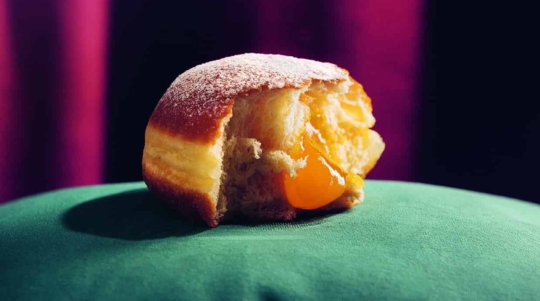 Nahaufnahme eines halb aufgegessenen, mit Gelee gefüllten Krapfen mit Puderzucker auf der Oberseite, aus dem leuchtend orangefarbene Füllung austritt und der auf einer grünen Fläche mit lila Hintergrund liegt.