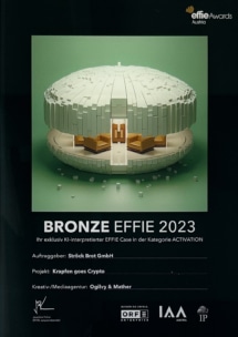 Die Plakette für den Effie 2023 in Bronze zeigt ein abstraktes, kreisförmiges, grün-weißes 3D-Gebäck mit einer goldenen Schleife. Sie würdigt die Ströck Brot GmbH für das Projekt "Krapfen goes Crypto" in der Kategorie "Activation".