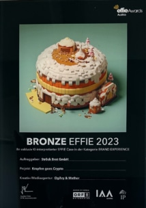 Ein Plakat für den Bronze-Effie 2023 zeigt eine Pixel-Art-Kuchen- und Hausszene auf schwarzem Hintergrund. Zu den Details gehören der Projekttitel "Krapfen goes Crypto" und die Kreativagentur Ogilvy & Mather mit dem Branding des Effie Awards Austria.
