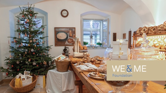 Eine gemütliche, weihnachtlich geschmückte Bäckerei mit einem festlichen Baum, Körben mit Brot und Gebäck auf einem Holztisch und dem Logo der WE & ME Foundation im Vordergrund.
