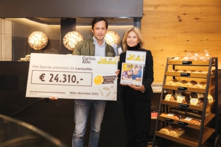 Ein Mann und eine Frau stehen drinnen und halten einen großen Spendenscheck über 24.310 € und ein Plakat in der Hand. Hinter ihnen stehen Regale mit Brot und eine holzgetäfelte Wand. Der Scheck ist adressiert für die Unterstützung von Lerncafés in Wien, November 2023.