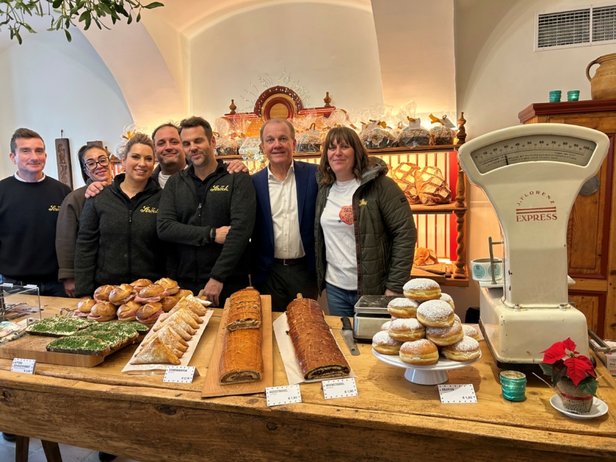 Sieben Personen stehen lächelnd hinter einer hölzernen Theke, die mit Gebäck, Brot und einer alten Waage gefüllt ist, in einer gemütlichen Bäckerei mit Regalen voller Backwaren im Hintergrund.