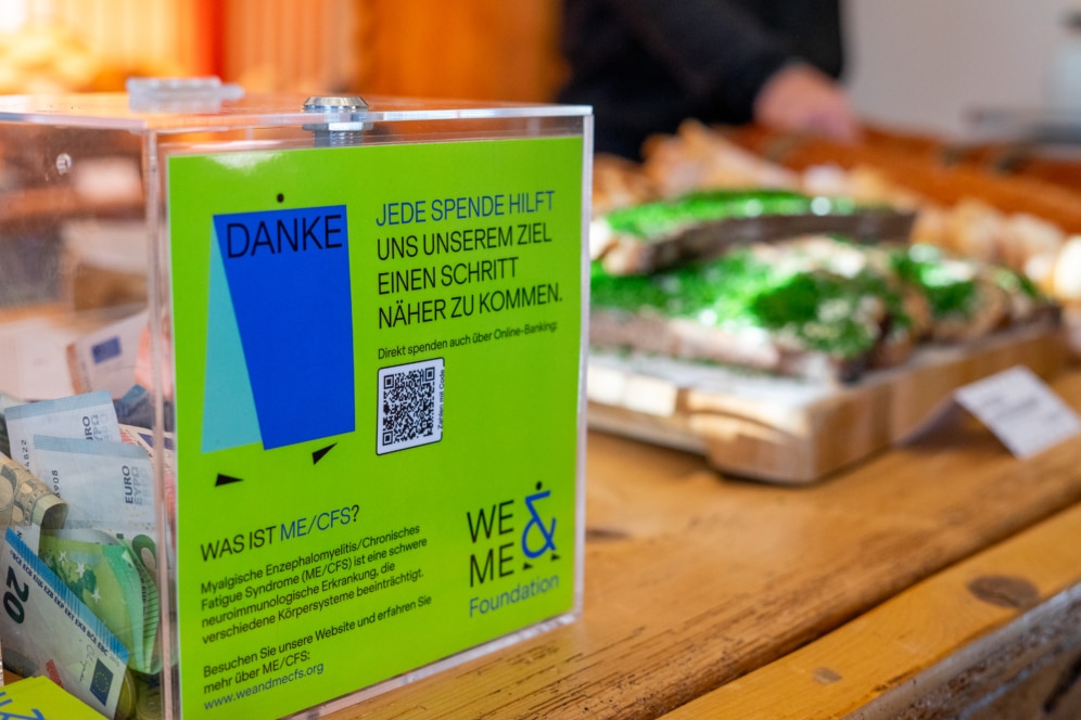 Auf einem Holztisch steht eine Spendenbox mit Euroscheinen und Münzen. Ein Schild auf Deutsch erklärt, wie Spenden das Bewusstsein für ME/CFS unterstützen, mit einem QR-Code und dem Logo der We & Me Foundation. Im Hintergrund sind Gebäckstücke zu sehen.