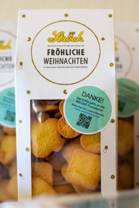 Eine durchsichtige Packung goldener Kekse mit der Aufschrift "Fröhliche Weihnachten" und einem runden grünen Aufkleber, der ein Dankeschön ausdrückt und auf eine Spende an die Stiftung WE4EINIGE hinweist.