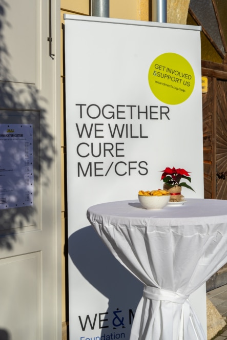 Ein runder Tisch mit Snacks und einem Weihnachtsstern steht vor einem Transparent mit der Aufschrift TOGETHER WE WILL CURE ME/CFS und einem Aufkleber mit der Aufschrift GET INVOLVED & SUPPORT US. Das Sonnenlicht erhellt die Szene.