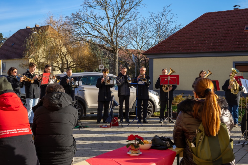 Eine Blaskapelle spielt im Freien in Wintermänteln vor einem auf Bänken sitzenden Publikum. Im Hintergrund sind Häuser und blattlose Bäume zu sehen, im Vordergrund ein roter Tisch mit einem Weihnachtsstern.
