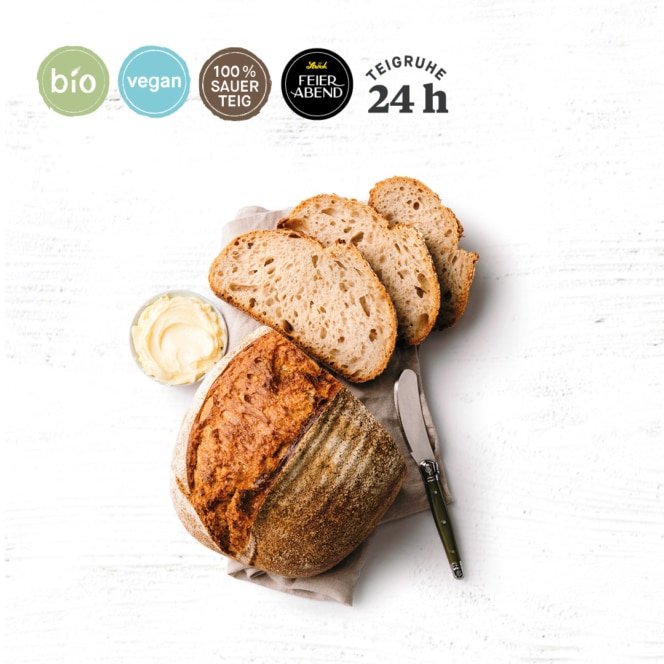Ein runder Laib rustikalen Brotes, teilweise aufgeschnitten, liegt auf einer Serviette mit einem Messer und einer kleinen Schale mit Butter. Vier Symbole oben zeigen an, dass das Brot biologisch, vegan, 100% Sauerteig und 24 Stunden geruht hat.
