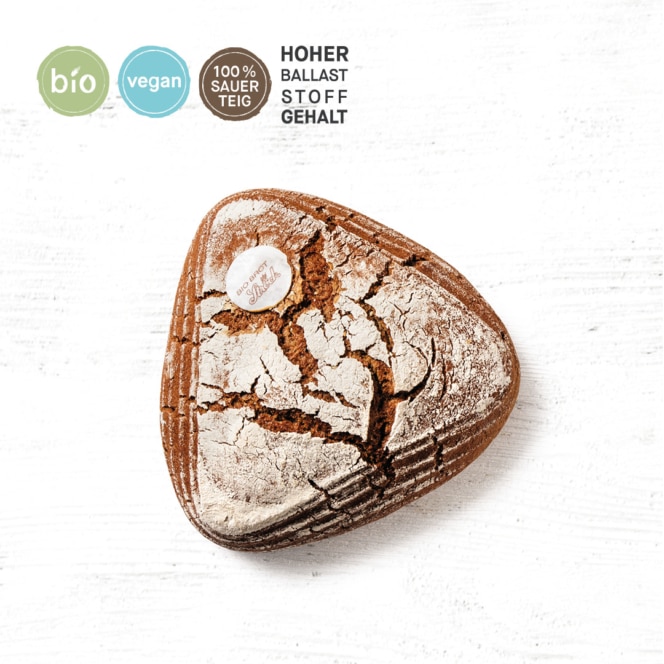 Ein dreieckiger Laib rustikales Schwarzbrot mit einer rissigen, mehlbestäubten Kruste liegt auf einer weißen Fläche. Kreisförmige Symbole kennzeichnen es als biologisch, vegan und 100% Sauerteig mit hohem Ballaststoffgehalt in deutscher Sprache.