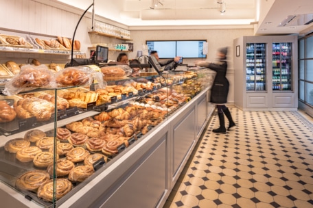 Eine Bäckerei mit Glasvitrinen, gefüllt mit verschiedenen Backwaren. Zwei Personen stehen hinter der Theke, und ein Kunde im Mantel steht davor. Im Hintergrund sind gekühlte Getränke zu sehen.