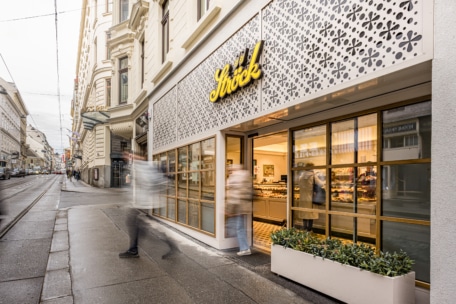 Eine moderne Bäckerei mit großen Fenstern und gemusterten Außenverkleidungen in einer Stadtstraße; verschwommene Gestalten gehen am Eingang vorbei, und im Inneren des gut beleuchteten Ladens ist Gebäck zu sehen.