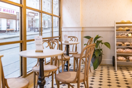 Eine gemütliche Bäckerei mit runden Tischen und Holzstühlen an großen Fenstern. In der Ecke steht eine Pflanze, und in den Regalen sind verschiedene Brotsorten ausgestellt. Der Boden hat ein schwarz-weißes geometrisches Fliesenmuster.
