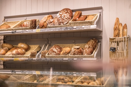 Auslage einer Bäckerei mit verschiedenen Brotsorten, darunter runde Brote und Baguettes, die in Weidenkörben auf Regalen vor einer weißen Wand angeordnet sind. Einige Brotsorten sind beschriftet, und im Vordergrund ist ein Teil einer Glastheke zu sehen.