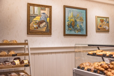 Das Innere einer Bäckerei zeigt Regale mit verschiedenen Brotsorten und Gebäck, und über den Backwaren hängen drei farbenfrohe Gemälde mit Lebensmitteln und Stillleben an einer hell gemusterten Wand.