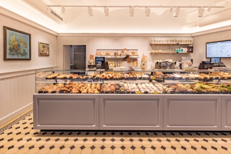 Eine helle Bäckerei mit einer Theke, die mit verschiedenen Backwaren, Brot und Donuts gefüllt ist. An den cremefarbenen Wänden hängt Kunst, und im Hintergrund stehen Regale mit Zubehör und Kaffeezubehör.