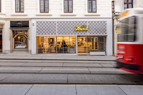 Eine Bäckerei mit großen Fenstern und einem Schild mit der Aufschrift "Ströck" ist von der anderen Seite einer Straße mit Straßenbahnschienen aus zu sehen, während eine rote Straßenbahn vorbeifährt, die aufgrund der Bewegung teilweise verschwommen ist.