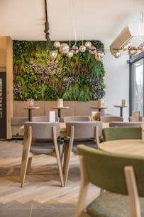 Moderne Café-Einrichtung mit Holztischen und gepolsterten Stühlen, mit einer grünen vertikalen Pflanzenwand als Hintergrund. Von der Decke hängen Hängeleuchten mit runden Glühbirnen, und durch große Fenster fällt Sonnenlicht ein.