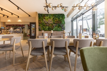 Modernes Café-Interieur mit hellen Holztischen und -stühlen, großen Fenstern auf der rechten Seite und einer grünen vertikalen Pflanzenwand im Hintergrund. Hängende Kugelleuchten spenden warmes Licht. Einige Speisekarten sind auf den Tischen platziert.