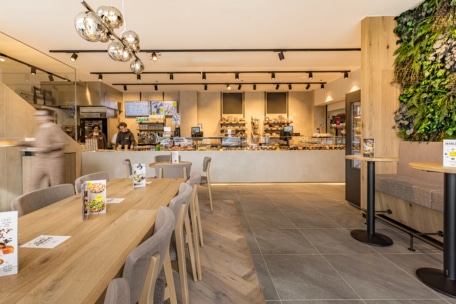 Ein modernes Bäckerei-Café mit Holztischen und -stühlen, einer Theke mit Backwaren, Mitarbeitern hinter der Theke und einer grünen Pflanzenwand auf der rechten Seite. Der Raum ist hell, stilvoll beleuchtet und neutral eingerichtet.