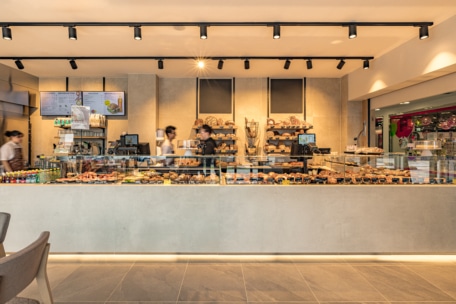 Eine moderne Bäckerei mit einer Glasvitrine, die mit verschiedenen Backwaren und Brot gefüllt ist. Drei Mitarbeiter arbeiten hinter der Theke, und an der Wand hinter ihnen sind Regale mit weiterem Brot zu sehen.