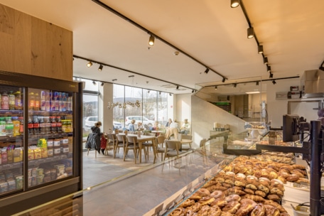 Eine moderne Bäckerei mit Glasvitrinen, die mit verschiedenen Backwaren und Broten gefüllt sind, einem Kühlschrank mit Getränken und Snacks und Tischen an großen Fenstern mit Tageslicht.