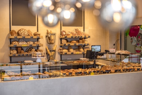 Eine Bäckerei mit Regalen, die mit verschiedenen Brotsorten gefüllt sind, und einer Glasvitrine, in der Gebäck, Croissants und Backwaren ausgestellt sind. Auf dem Tresen steht eine Registrierkasse. Die Beleuchtung ist warm.