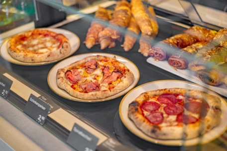 Hinter einer Glastheke in einer Bäckerei oder einem Café werden drei Pizzen mit verschiedenen Belägen auf Tellern neben verschiedenen Gebäcksorten ausgestellt. Die Preisschilder sind vor jedem Produkt sichtbar.